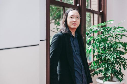 A portrait of Audrey Tang in semi-formal clothing (a dark blue shirt and near-black suit), leaning on a window frame and looking relaxed. Tang is standing next to a dark green leafy plant, and the window is painted dark brown at the corner of a room with whitewashed walls, a mix between modernity, tradition, and nature. Behind Tang, outside, there is more foliage.